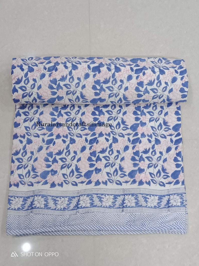 Op de afbeelding: Een witte stof met een blauwe en roze bloemenprint. De stof heeft een blauwe rand met een wit en blauw geometrisch patroon.