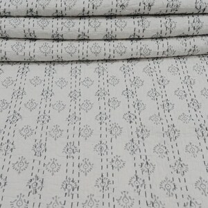 May include: A white cotton kantha quilt with a grey floral pattern. The quilt is folded in a stack and is sitting on a woven bamboo mat.