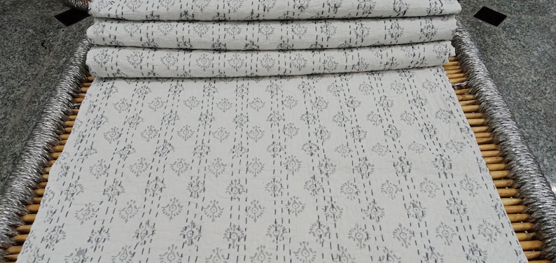 May include: A white cotton kantha quilt with a grey floral pattern. The quilt is folded in a stack and is sitting on a woven bamboo mat.