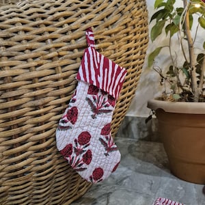 Puede incluir: Un pequeño calcetín navideño acolchado con fondo blanco y estampado floral rojo. La parte superior del calcetín presenta un diseño de rayas de cebra rojas y blancas. Se adjunta un lazo de tela roja para colgar.