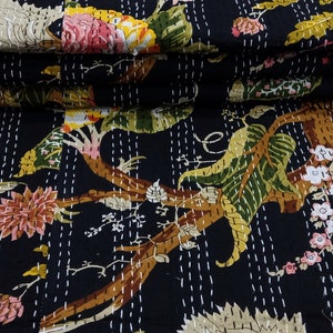 May include: A black kantha quilt with a floral pattern featuring trees, leaves, and flowers in shades of pink, green, yellow, and brown. The quilt is folded in a stack on a woven bamboo surface.