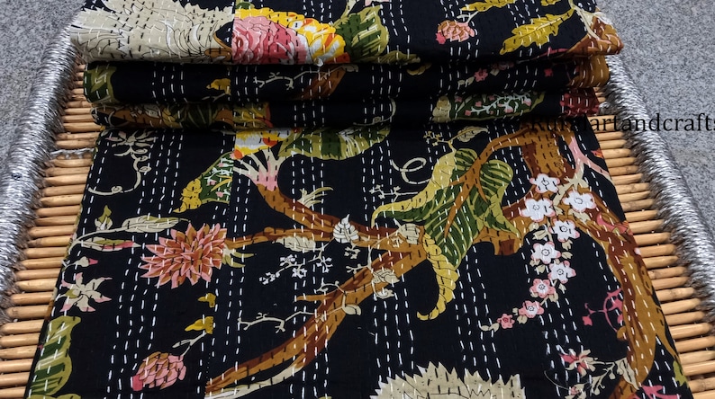 May include: A black kantha quilt with a floral pattern featuring trees, leaves, and flowers in shades of pink, green, yellow, and brown. The quilt is folded in a stack on a woven bamboo surface.