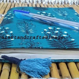 May include: A teal blue notebook with a floral pattern and a black tassel. The notebook is closed and a blue pen is lying on top of it. The notebook is sitting on a woven bamboo surface.