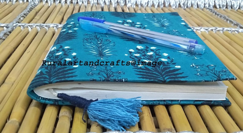 May include: A teal blue notebook with a floral pattern and a black tassel. The notebook is closed and a blue pen is lying on top of it. The notebook is sitting on a woven bamboo surface.