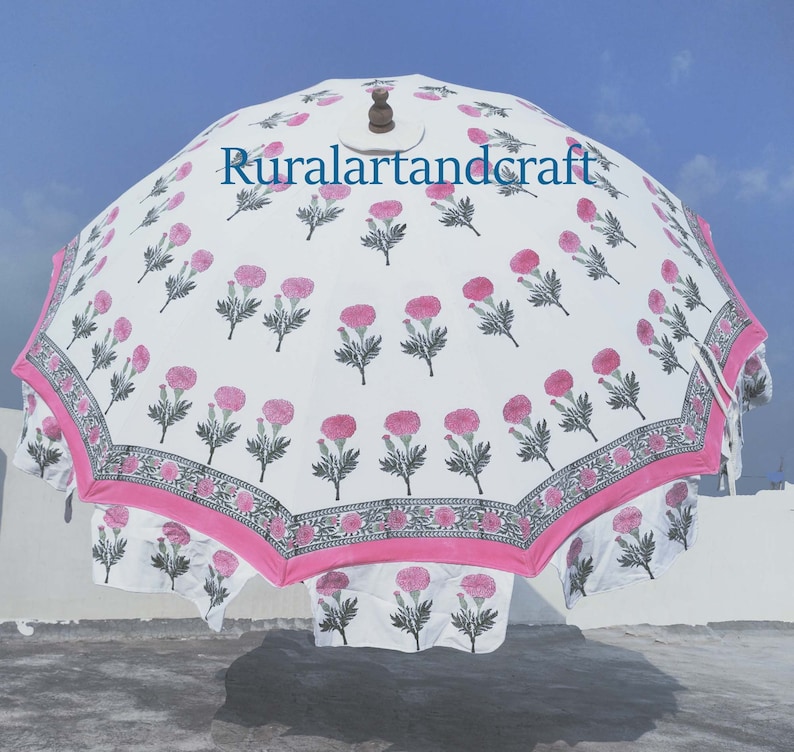 Op de afbeelding: Een witte en roze outdoor parasol met een bloemenpatroon en een houten handvat. De parasol heeft een roze rand en een herhalend patroon van roze bloemen met groene stelen en bladeren. De tekst "Ruralartandcraft" is op de parasol gedrukt.