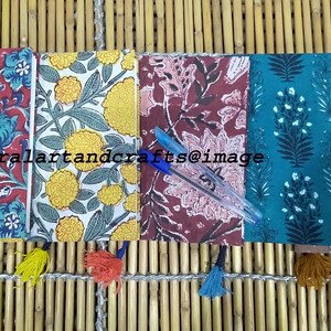 May include: Five small notebooks with floral patterns and tassels. The notebooks are arranged in a row on a woven bamboo surface. The notebooks are red, yellow, pink, teal, and beige.