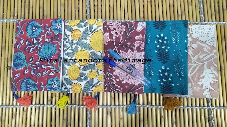 May include: Five small notebooks with floral patterns and tassels. The notebooks are arranged in a row on a woven bamboo surface. The notebooks are red, yellow, pink, teal, and beige.