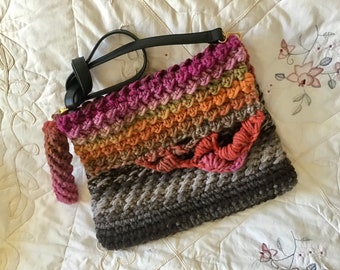 May include: A crocheted handbag with a black leather strap and a multi-colored ombre design. The bag is made with a chunky yarn in shades of pink, orange, brown, and gray.