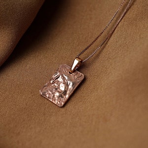 May include: A delicate gold necklace with a textured rectangular pendant. The pendant has a rough, organic look, like a piece of rock.
