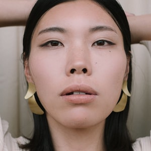 Puede incluir: Una mujer con cabello oscuro que lleva una blusa blanca texturizada y pendientes de hoja de oro.