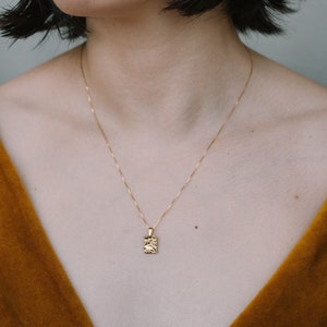 Puede incluir: Una mujer con el pelo negro corto lleva un collar de oro con un colgante cuadrado texturizado. Lleva una blusa de terciopelo marrón.