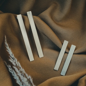 Puede incluir: Cuatro pendientes rectangulares de plata sobre un fondo de tela marrón. Los pendientes están dispuestos en dos pares, con el par más grande en la parte superior y el par más pequeño en la parte inferior.