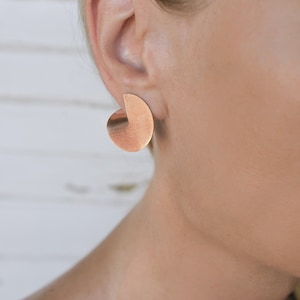 May include: A pair of gold-toned geometric earrings with a semi-circle shape. The earrings are made of metal and have a brushed finish.
