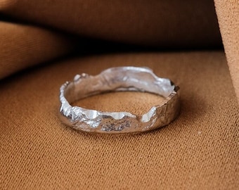 May include: A silver ring with a textured, irregular band. The ring is resting on a brown fabric surface.