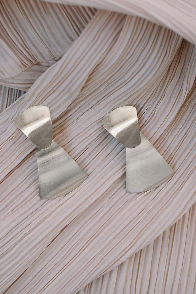 May include: A pair of silver geometric earrings with a unique, asymmetrical design. The earrings are made of a smooth, metallic material and have a modern, minimalist aesthetic.