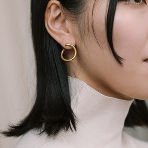 May include: A close-up shot of a person wearing a gold-colored, open-circle earring. The earring is a simple, modern design. The person is wearing a white turtleneck sweater and has dark hair.