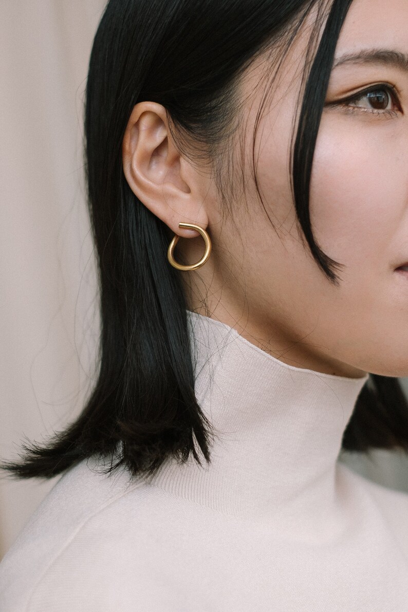 May include: A close-up shot of a person wearing a gold-colored, open-circle earring. The earring is a simple, modern design. The person is wearing a white turtleneck sweater and has dark hair.