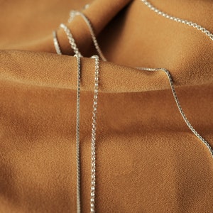 May include: Two silver chains lay on a brown fabric background. The chains are made of small, linked circles.