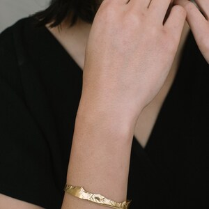 May include: A gold-toned bracelet with a textured, irregular shape. The bracelet is worn on a person's wrist.