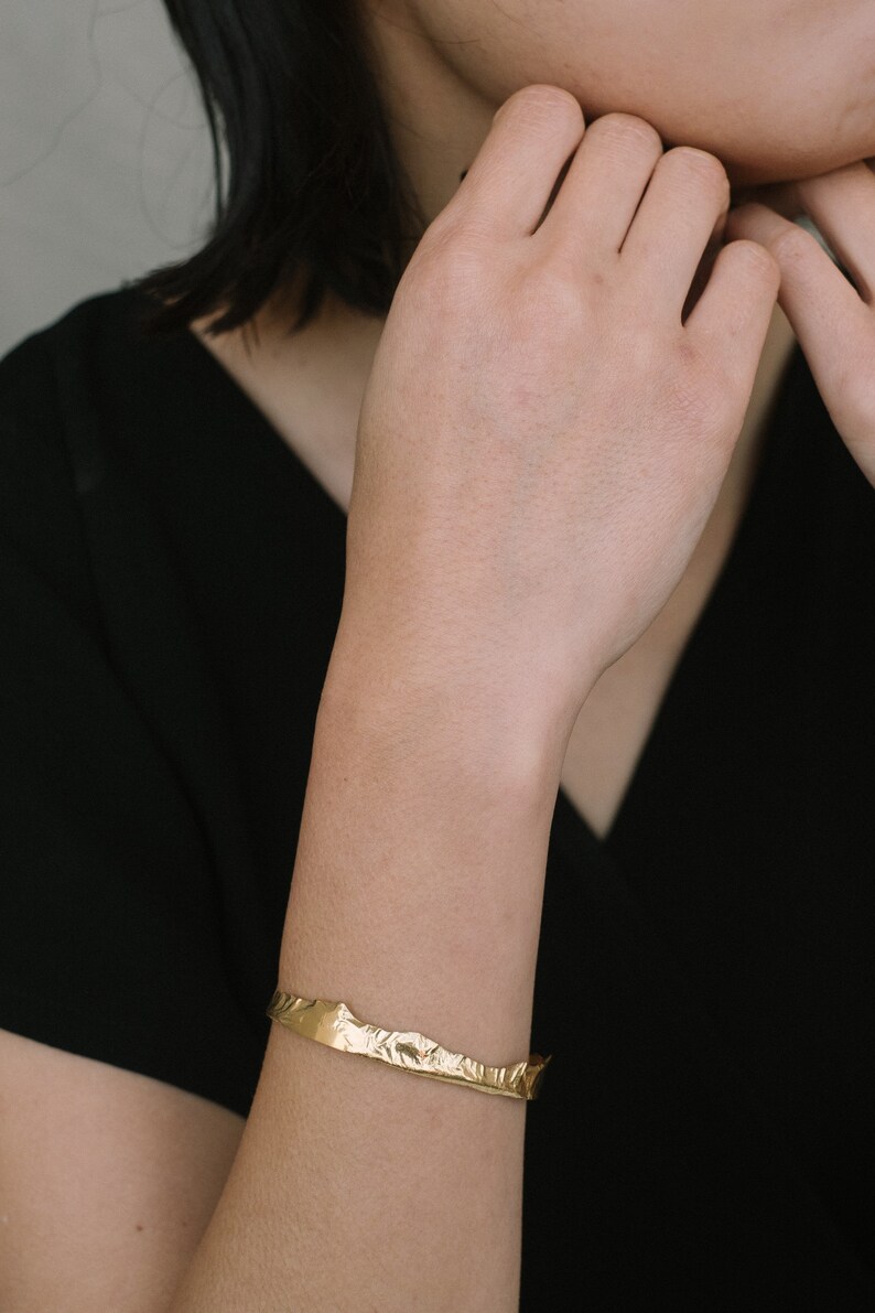 May include: A gold-toned bracelet with a textured, irregular shape. The bracelet is worn on a person's wrist.