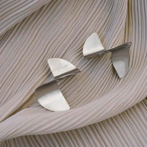 May include: Two silver abstract earrings with a twisted design, displayed on a beige pleated fabric background.