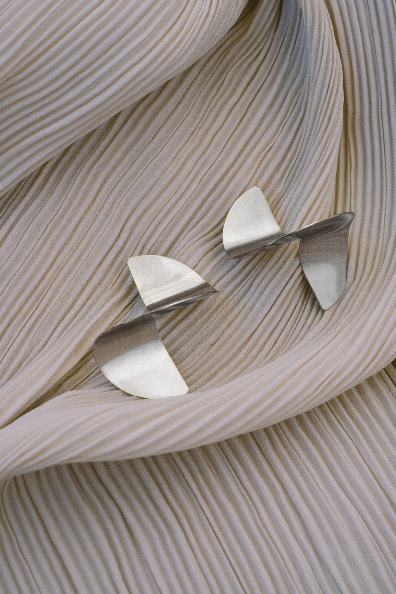 May include: Two silver abstract earrings with a twisted design, displayed on a beige pleated fabric background.