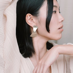 May include: A woman with short black hair wearing a light beige top with a v-neckline and large gold geometric earrings.