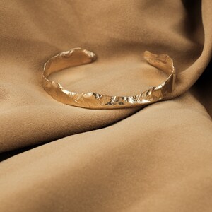 May include: A gold-toned, textured metal bracelet with a rough, organic design. The bracelet is lying on a brown fabric background.