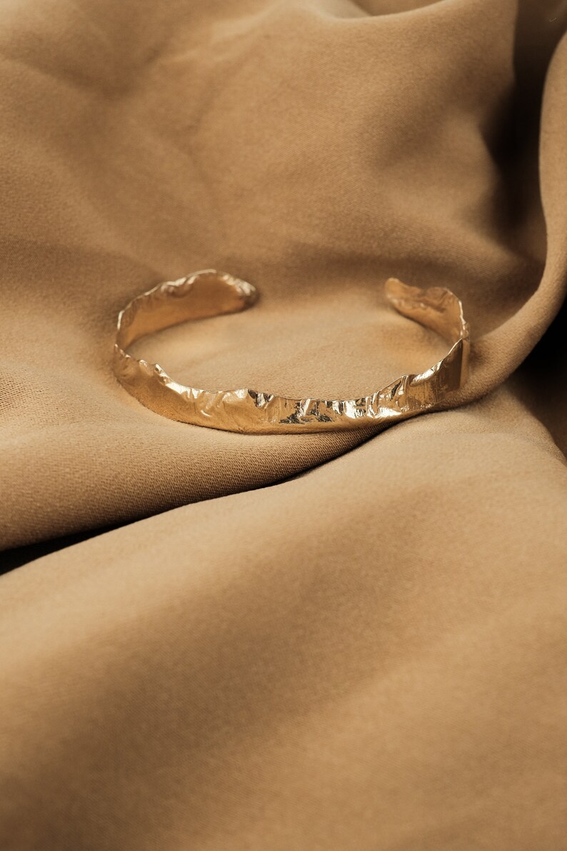 May include: A gold-toned, textured metal bracelet with a rough, organic design. The bracelet is lying on a brown fabric background.