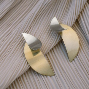 May include: A pair of gold and silver earrings with a crescent moon shape. The earrings are on a beige fabric background.