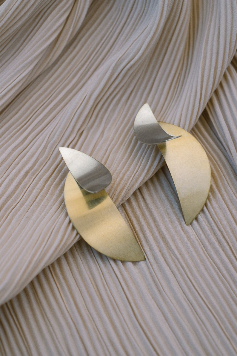 May include: A pair of gold and silver earrings with a crescent moon shape. The earrings are on a beige fabric background.