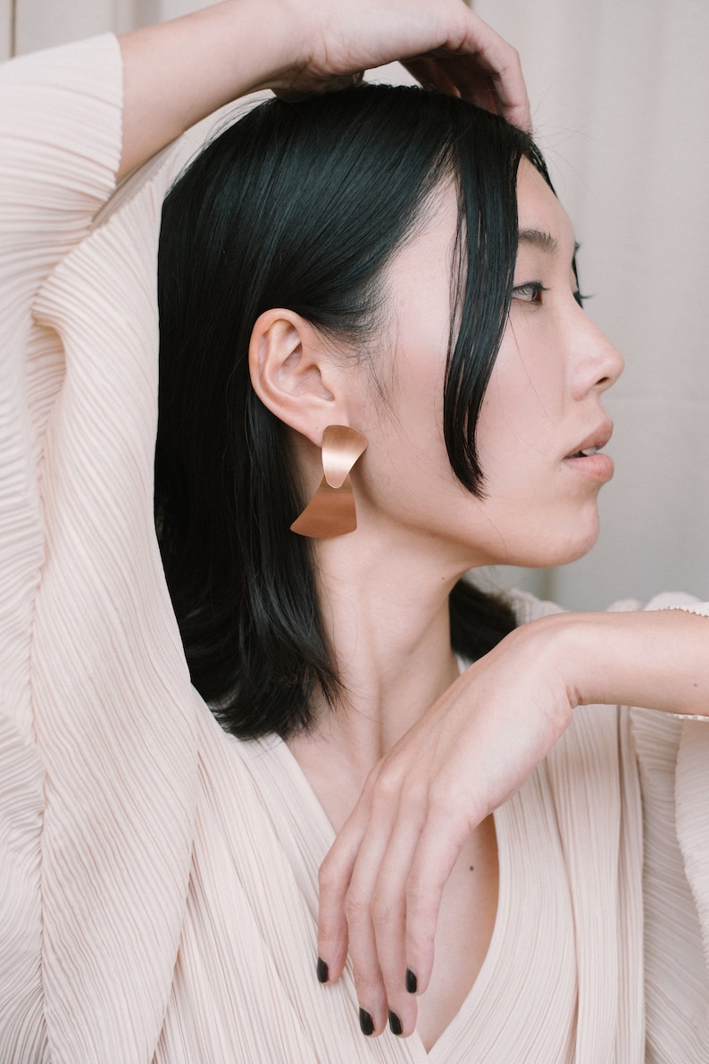 May include: A woman with short black hair wearing a light beige top with a v-neckline. She is wearing large gold earrings with a geometric design.