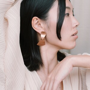 Puede incluir: Una mujer con el pelo negro corto lleva una blusa beige claro con escote en V. Lleva unos pendientes grandes de oro con un dise&ntilde;o geom&eacute;trico.