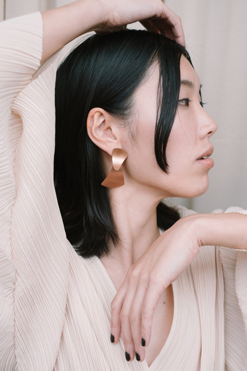 Puede incluir: Una mujer con el pelo negro corto lleva una blusa beige claro con escote en V. Lleva unos pendientes grandes de oro con un dise&ntilde;o geom&eacute;trico.