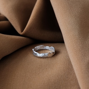 May include: A silver ring with a textured, irregular, and slightly jagged edge. The ring is lying on a brown fabric surface.