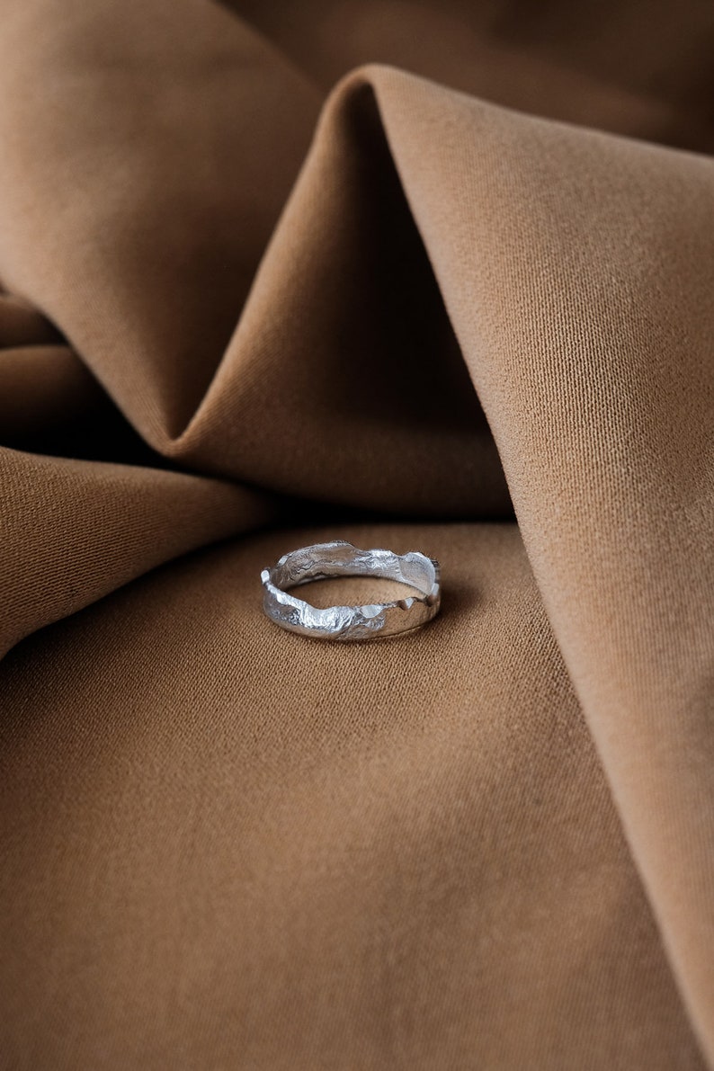 May include: A silver ring with a textured, irregular, and slightly jagged edge. The ring is lying on a brown fabric surface.