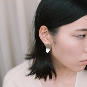 May include: A woman with short black hair wearing a pair of gold geometric earrings. The earrings are shaped like a triangle with rounded edges.