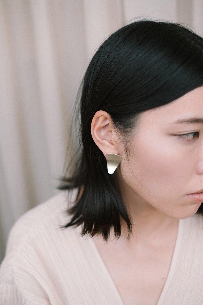 May include: A woman with short black hair wearing a pair of gold geometric earrings. The earrings are shaped like a triangle with rounded edges.