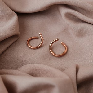 May include: A pair of copper-colored ear cuffs. Each cuff has a curved, open design. The earrings are displayed on a draped, neutral-toned fabric, creating a minimalist aesthetic. The warm tones of the earrings contrast with the fabric.
