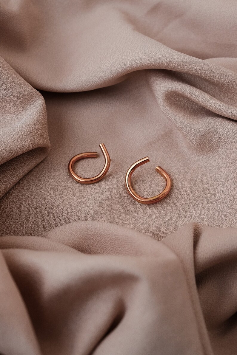 May include: A pair of copper-colored ear cuffs. Each cuff has a curved, open design. The earrings are displayed on a draped, neutral-toned fabric, creating a minimalist aesthetic. The warm tones of the earrings contrast with the fabric.