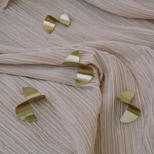 May include: Four gold-coloured metal shapes in the form of a crescent moon, arranged in a pattern on a light pink fabric background.
