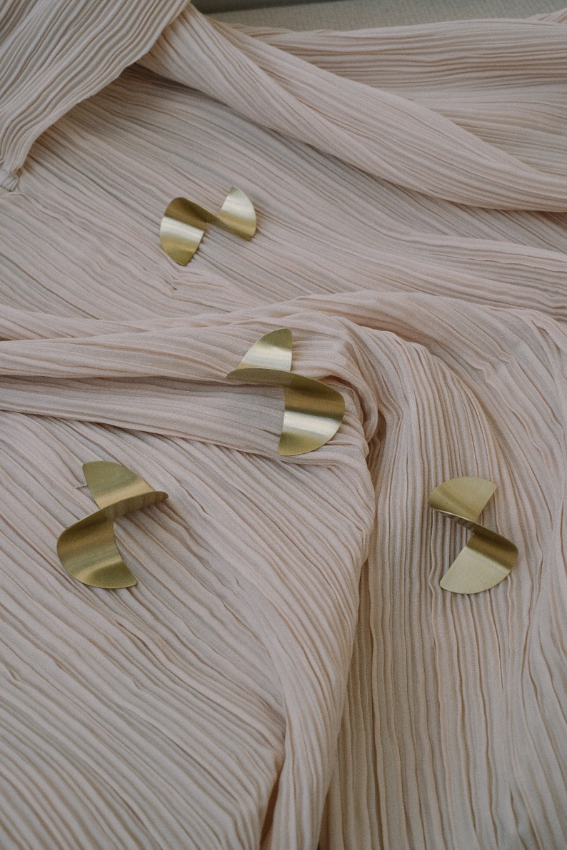 May include: Four gold-coloured metal shapes in the form of a crescent moon, arranged in a pattern on a light pink fabric background.