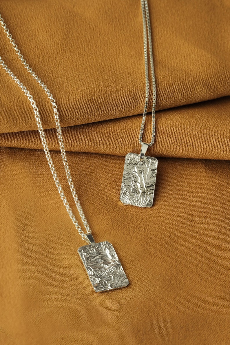 May include: Two silver necklaces with textured rectangular pendants on a brown fabric background.