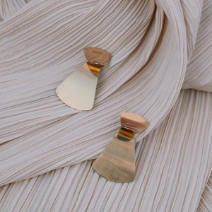 May include: A pair of gold-toned earrings with a geometric, fan-shaped design. The earrings are displayed on a cream-coloured, pleated fabric.