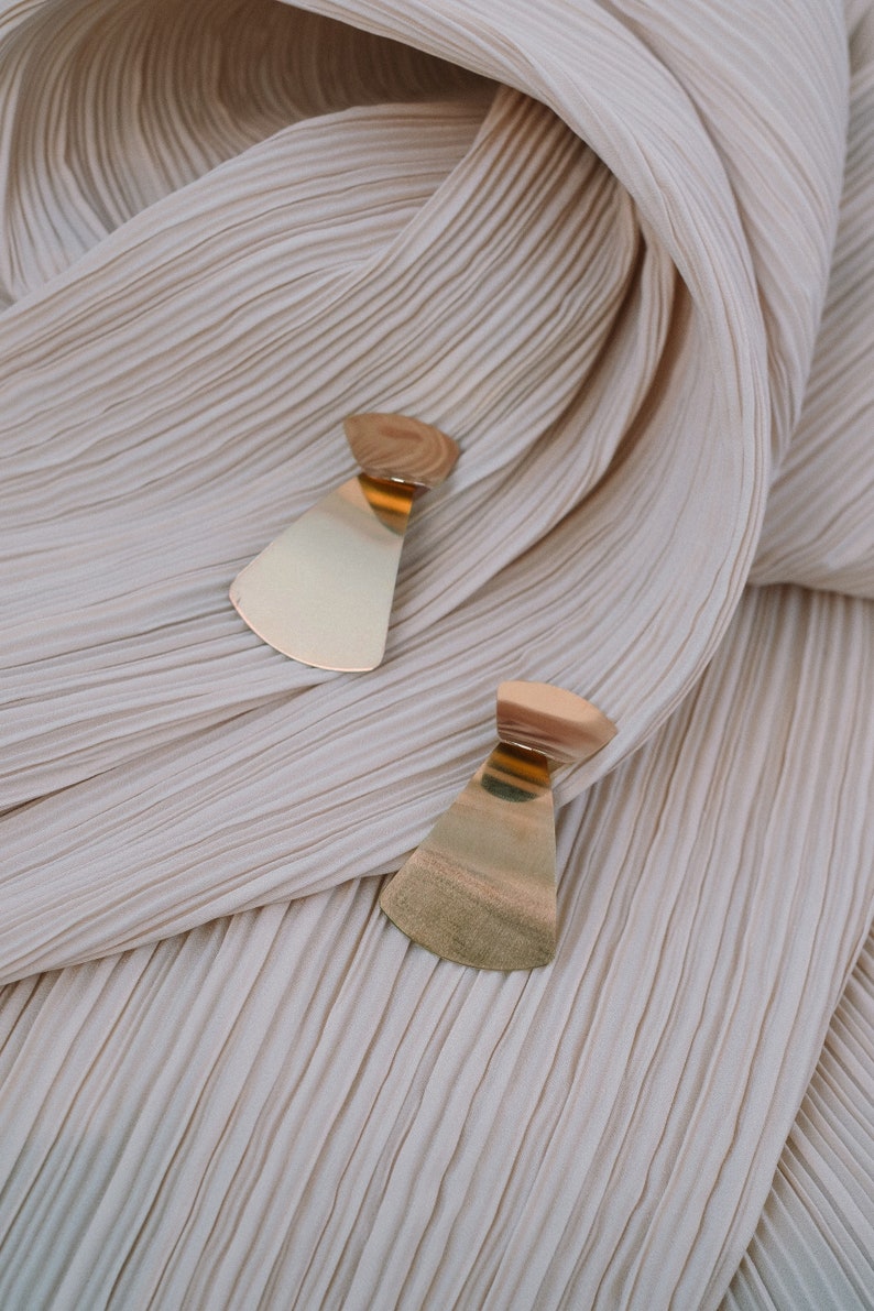 May include: A pair of gold-toned earrings with a geometric, fan-shaped design. The earrings are displayed on a cream-coloured, pleated fabric.