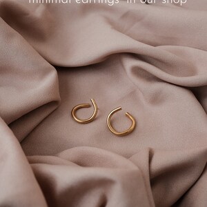 May include: Two gold-colored, minimalist earrings with a curved design, resting on a draped, light brown fabric. The text in the image reads: "find thinner models/other sizes in the section 'minimal earrings' in our shop."