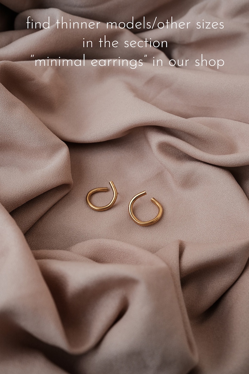 May include: Two gold-colored, minimalist earrings with a curved design, resting on a draped, light brown fabric. The text in the image reads: "find thinner models/other sizes in the section 'minimal earrings' in our shop."