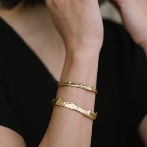 May include: Two gold-toned bracelets with a textured, mountain-like design. The bracelets are stacked on a person's wrist.