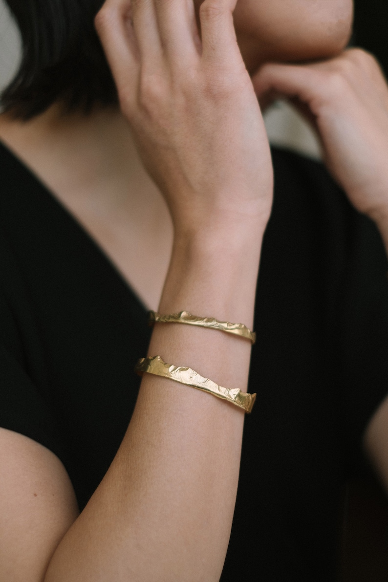 May include: Two gold-toned bracelets with a textured, mountain-like design. The bracelets are stacked on a person's wrist.