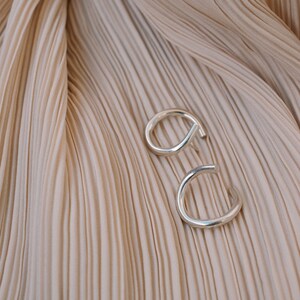 May include: Two silver-colored, open-ended hoop earrings. The earrings are set against a backdrop of pleated, beige fabric. The earrings have a simple, modern design, with a curved shape that wraps around the earlobe.